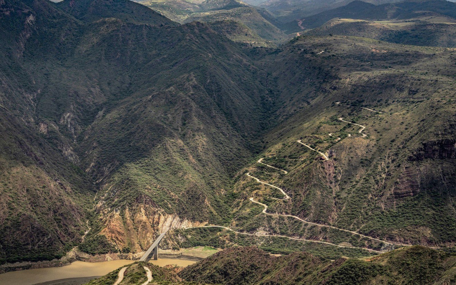 A winding switchback road climbs steep mountainous terrain above a bridge crossing a muddy river