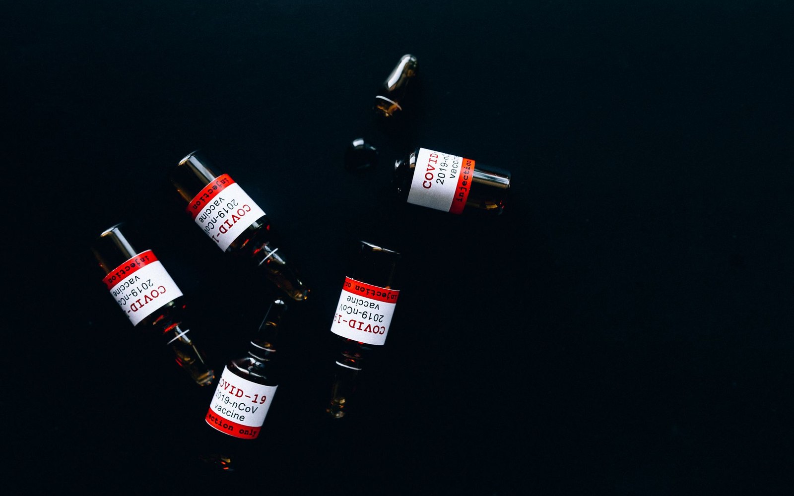 Small glass vials labeled covid-19 vaccine scattered on a dark reflective surface