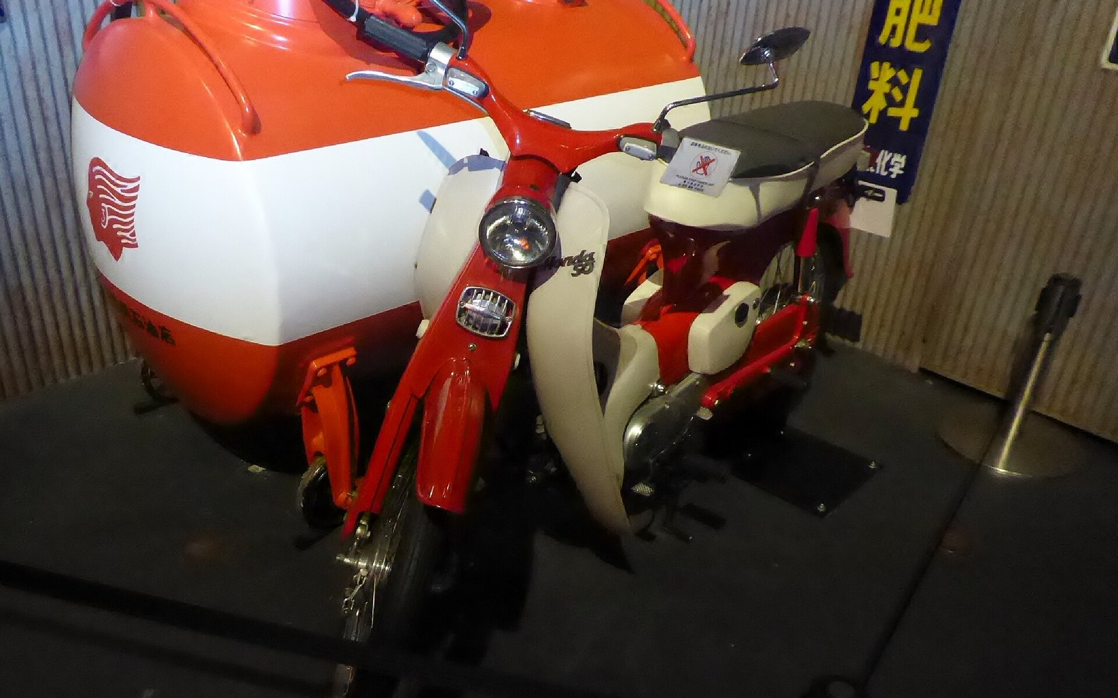 Vintage red and white Honda Super Cub scooter displayed beside a large tanker-shaped exhibit