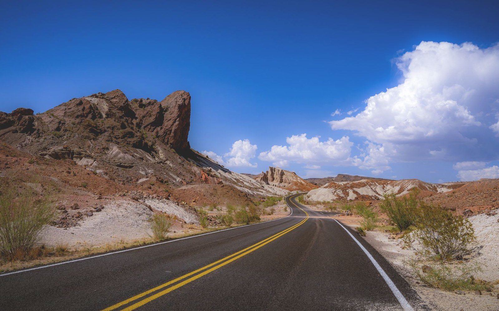 A winding desert road curves past jagged red rock formations under a blue sky
