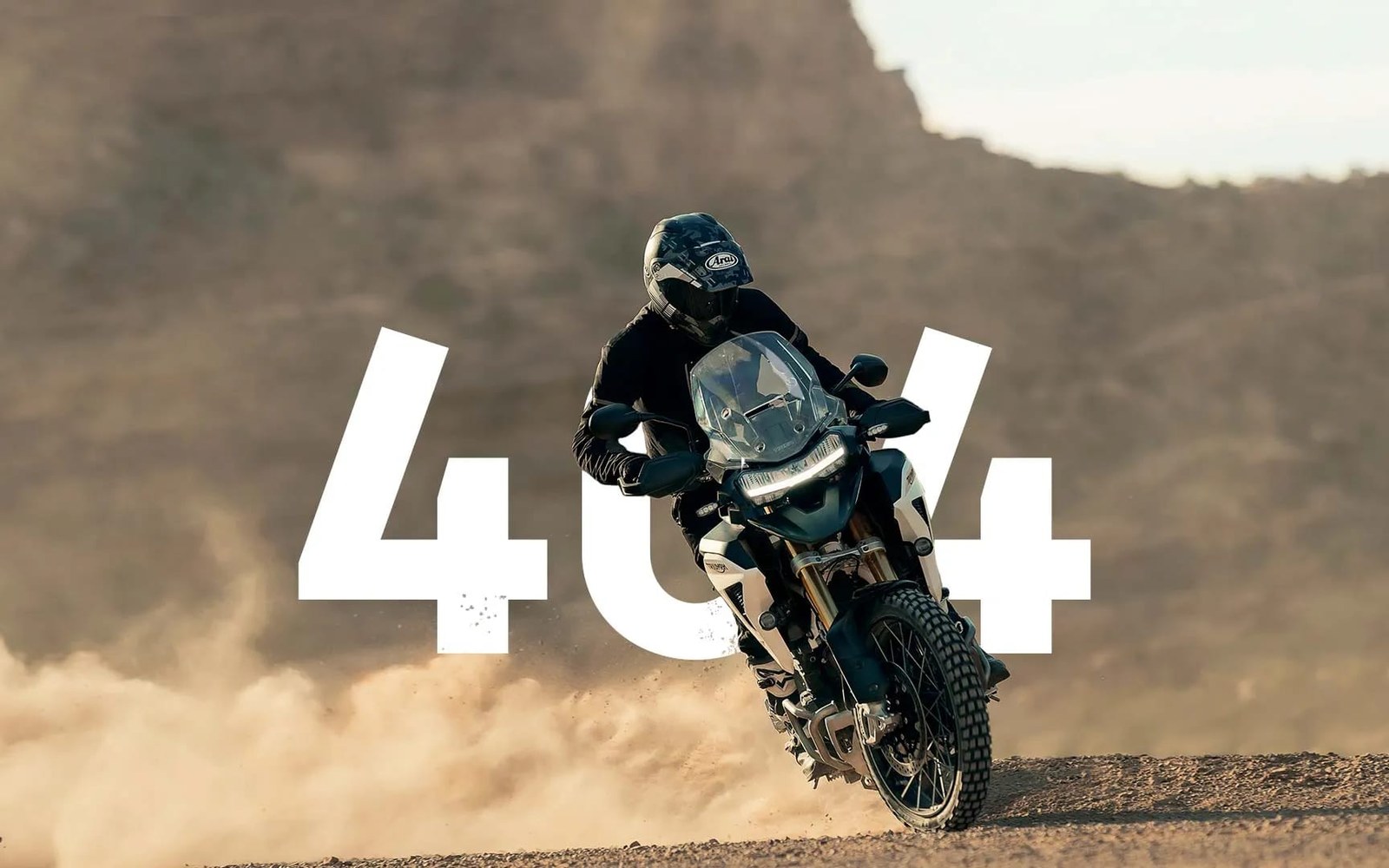 Rider kicks up dust on an adventure motorcycle with a "404" overlay on a desert track