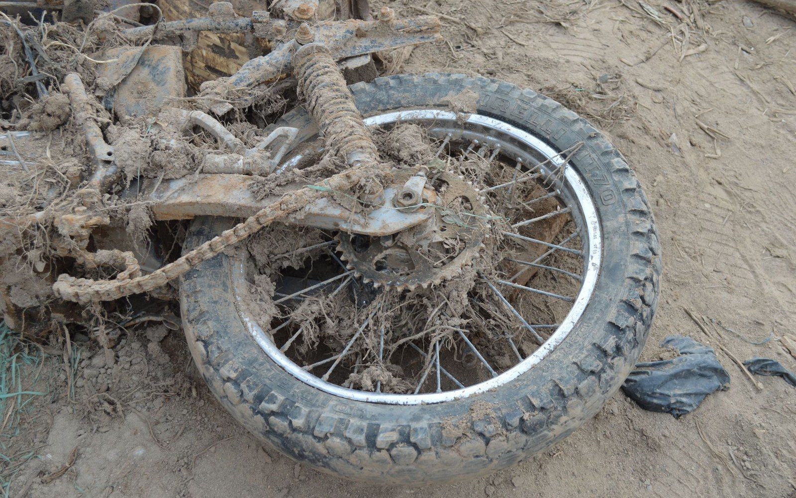 Mud-caked motorcycle rear wheel and chain lying on sandy ground
