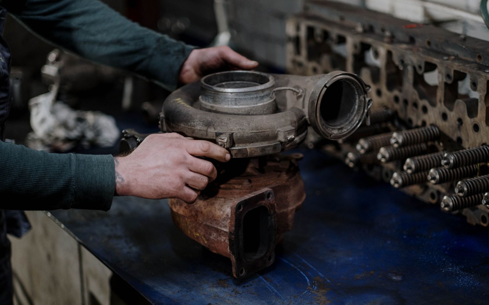 Adult mechanic's hands working on an opened parallel-twin engine on a workshop bench, overhead fluorescent light, tools laid on a cloth, oil on metal
